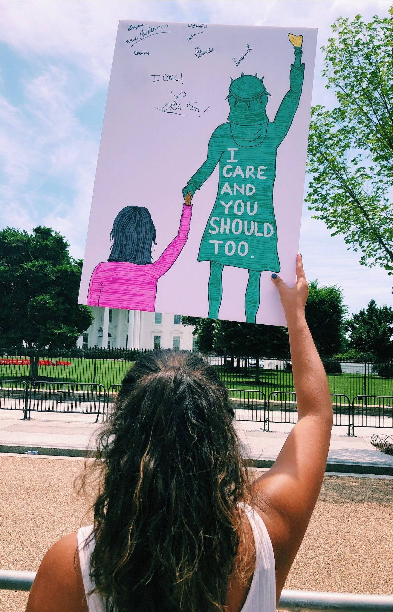 Image of woman holding a sign in front of the U.S. White House. The sign shows the Statue of Liberty, wearing a jacket that says “I care and you should too,” while holding the hand of an immigrant child.